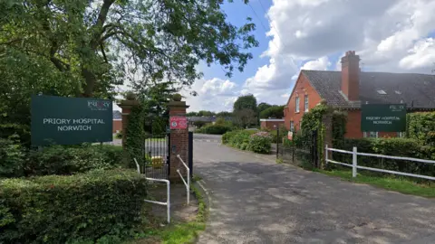 Google The entrance gate to Priory Hospital Norwich on a clear day with blue skies. A red brick building is on one side of the road with trees on the other.