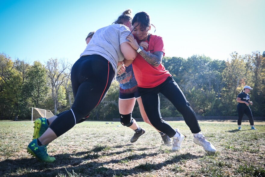 Three women are shown colliding on a grassy field. Two at right are pushing against one at left.