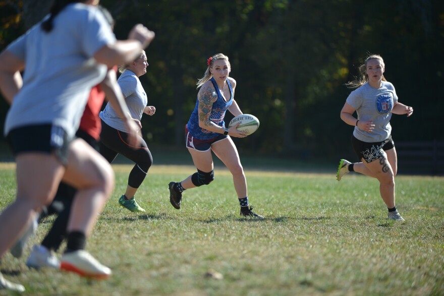 Several women run on a grassy field. One in the middle is carrying a rugby ball and looking like she is going to toss it.