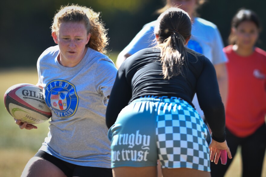 A woman carrying a rugby ball in her right hand runs toward a player  about to push the player out of the way with her left hand. The other player's shorts read 