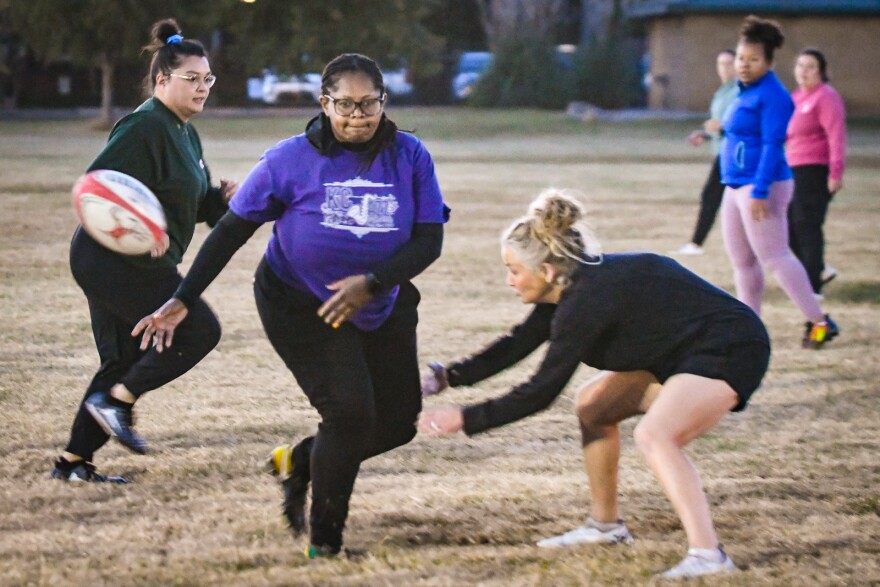 One woman in the center of the frame wears a purple T-shirt over a black long-sleeve shirt. She appears to be running and tossing a rugby ball while other players swarm around her.