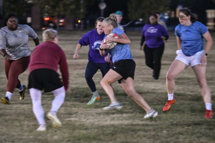 Women are shown outside on a grassy field. One in the middle of the photo is carrying a rugby ball and running  between the other women who appear to be trying to tackle her.