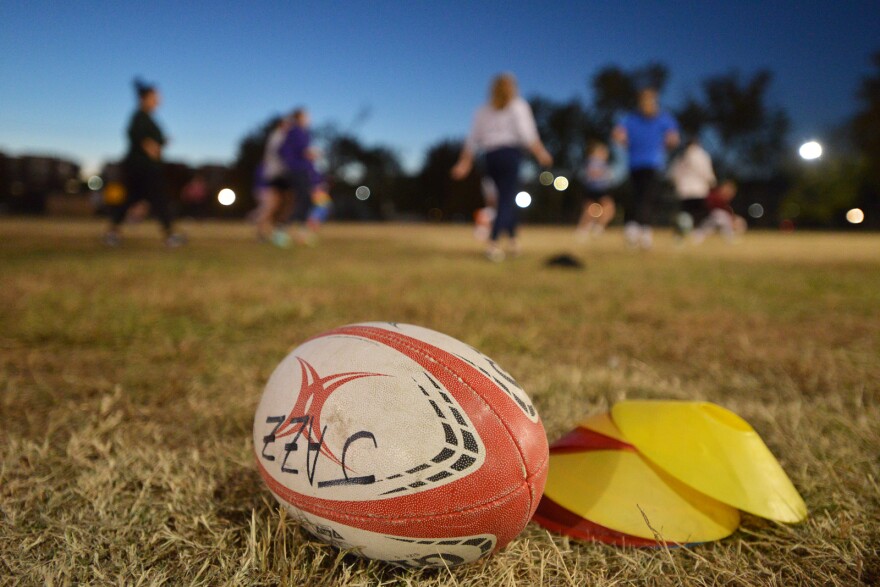 Wide angle shot of a rugby in the foreground ball sitting on a field in waning daylight. Behind it are out-of-focus people running  in a loose group. Written on the ball is the word 