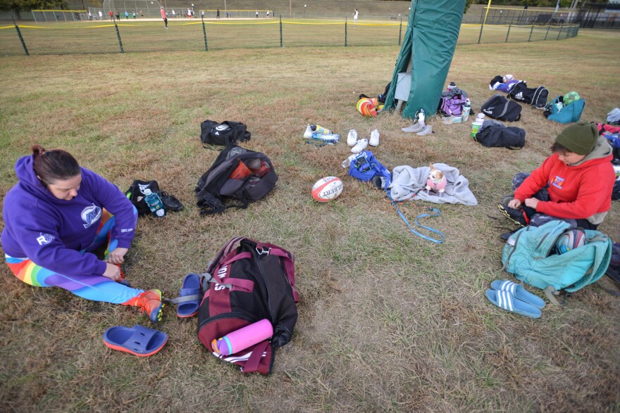 Two women are shown sitting on a grassy field. They are sitting surrounded by small duffel bags and other clothing. A rugby ball is on the ground between them.