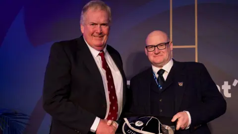 Scottish Rugby Two men in formal attire are standing and shaking hands. The man on the left has light brown hair and is wearing a dark navy suit jacket, a white shirt, and a maroon patterned tie. The man on the right is bald, has glasses, and is wearing a navy blue suit jacket.