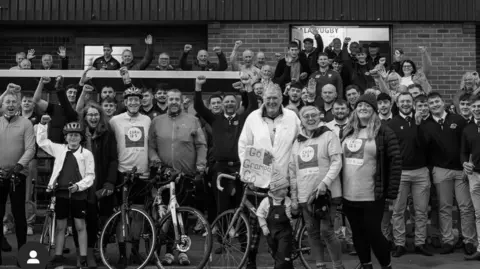 Gala Rugby Full black and white outdoor shot of a large group of people, many on bicycles, in front of a grandstand at a sports venue. The grandstand is a multi-tiered structure with rows of light beige/cream-colored seats.