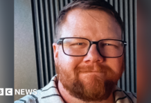 Mark Orpin in a family photo wearing a grey polo top with black stripes. He has shot cropped brown hair, an ginger beard beneath black rimmed glasses. He has closed mouth smile and has a kind face