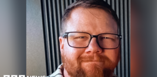 Mark Orpin in a family photo wearing a grey polo top with black stripes. He has shot cropped brown hair, an ginger beard beneath black rimmed glasses. He has closed mouth smile and has a kind face