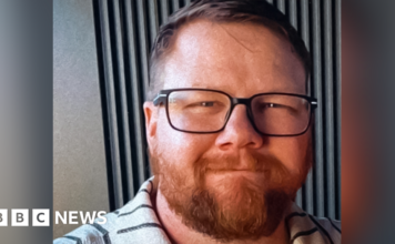 Mark Orpin in a family photo wearing a grey polo top with black stripes. He has shot cropped brown hair, an ginger beard beneath black rimmed glasses. He has closed mouth smile and has a kind face