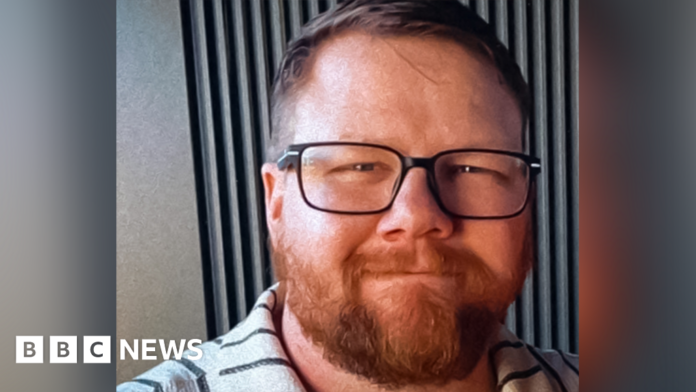 Mark Orpin in a family photo wearing a grey polo top with black stripes. He has shot cropped brown hair, an ginger beard beneath black rimmed glasses. He has closed mouth smile and has a kind face