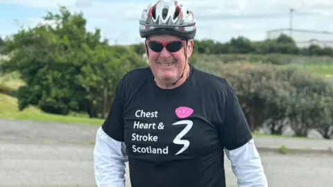 Scottish Rugby Ken MacAulay is wearing a black t-shirt  over a white cycling shirt. The shirt has the words 