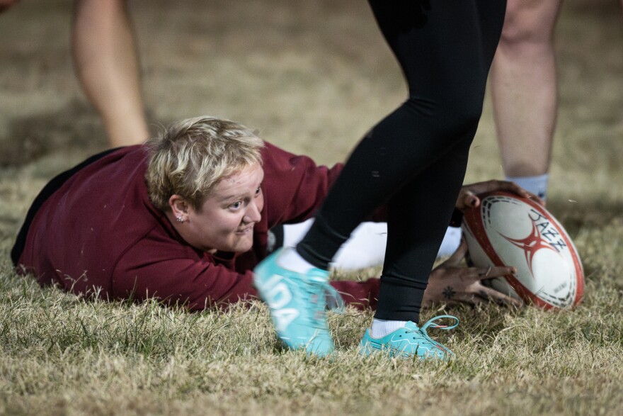 A close shot of a woman wearing a maroon sweatshirt. She is lying on the ground with outstretched hand placing a rugby ball on the ground. The legs of players can be seen around her.