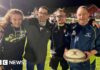 Four men embrace on a rugby pitch with the man on the right holding a rugby ball. The man on the left has long hair and is wearing a navy hoodie. Next to him is a man in a grey coat and glasses. Next to him is a man in glasses, a blue tracksuit top and shirt. To the right is a man in full navy tracksuit. They are all smiling under the floodlights.