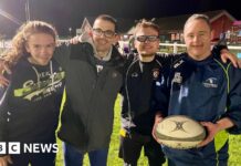 Four men embrace on a rugby pitch with the man on the right holding a rugby ball. The man on the left has long hair and is wearing a navy hoodie. Next to him is a man in a grey coat and glasses. Next to him is a man in glasses, a blue tracksuit top and shirt. To the right is a man in full navy tracksuit. They are all smiling under the floodlights.