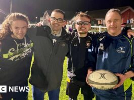 Four men embrace on a rugby pitch with the man on the right holding a rugby ball. The man on the left has long hair and is wearing a navy hoodie. Next to him is a man in a grey coat and glasses. Next to him is a man in glasses, a blue tracksuit top and shirt. To the right is a man in full navy tracksuit. They are all smiling under the floodlights.