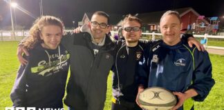 Four men embrace on a rugby pitch with the man on the right holding a rugby ball. The man on the left has long hair and is wearing a navy hoodie. Next to him is a man in a grey coat and glasses. Next to him is a man in glasses, a blue tracksuit top and shirt. To the right is a man in full navy tracksuit. They are all smiling under the floodlights.