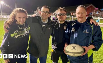 Four men embrace on a rugby pitch with the man on the right holding a rugby ball. The man on the left has long hair and is wearing a navy hoodie. Next to him is a man in a grey coat and glasses. Next to him is a man in glasses, a blue tracksuit top and shirt. To the right is a man in full navy tracksuit. They are all smiling under the floodlights.