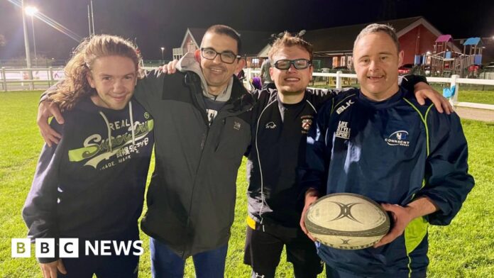 Four men embrace on a rugby pitch with the man on the right holding a rugby ball. The man on the left has long hair and is wearing a navy hoodie. Next to him is a man in a grey coat and glasses. Next to him is a man in glasses, a blue tracksuit top and shirt. To the right is a man in full navy tracksuit. They are all smiling under the floodlights.