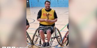 Patrick Murphy, sat in his wheelchair and holding a rugby league trophy in a gymnasium.