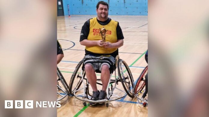 Patrick Murphy, sat in his wheelchair and holding a rugby league trophy in a gymnasium.