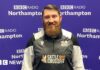 A man with short brown hair brushed to the left with a big beard stands in front of the purple BBC Radio Northampton branded wall smiling at the camera. He is wearing a black, white and grey zipped top with "The Man Cave" written above the left side of his chest.