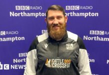 A man with short brown hair brushed to the left with a big beard stands in front of the purple BBC Radio Northampton branded wall smiling at the camera. He is wearing a black, white and grey zipped top with "The Man Cave" written above the left side of his chest.