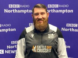 A man with short brown hair brushed to the left with a big beard stands in front of the purple BBC Radio Northampton branded wall smiling at the camera. He is wearing a black, white and grey zipped top with "The Man Cave" written above the left side of his chest.