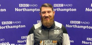 A man with short brown hair brushed to the left with a big beard stands in front of the purple BBC Radio Northampton branded wall smiling at the camera. He is wearing a black, white and grey zipped top with "The Man Cave" written above the left side of his chest.