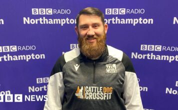 A man with short brown hair brushed to the left with a big beard stands in front of the purple BBC Radio Northampton branded wall smiling at the camera. He is wearing a black, white and grey zipped top with "The Man Cave" written above the left side of his chest.