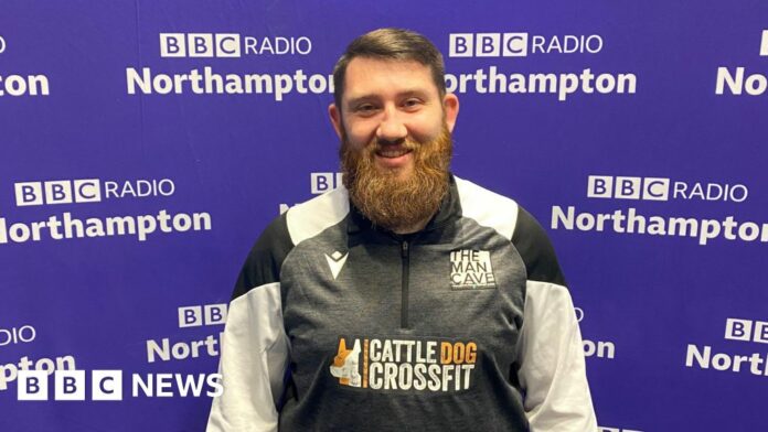 A man with short brown hair brushed to the left with a big beard stands in front of the purple BBC Radio Northampton branded wall smiling at the camera. He is wearing a black, white and grey zipped top with "The Man Cave" written above the left side of his chest.