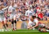 A woman in white England kit looks thrilled as she scores a rugby try with three other England players behind her looking equally happy