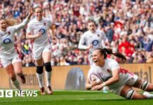 A woman in white England kit looks thrilled as she scores a rugby try with three other England players behind her looking equally happy