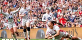 A woman in white England kit looks thrilled as she scores a rugby try with three other England players behind her looking equally happy