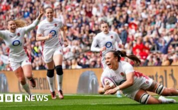 A woman in white England kit looks thrilled as she scores a rugby try with three other England players behind her looking equally happy