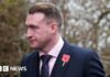 A man in a suit and tie wearing a poppy looks off to the left with trees in the background