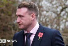 A man in a suit and tie wearing a poppy looks off to the left with trees in the background