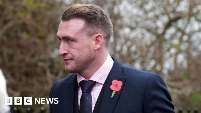 A man in a suit and tie wearing a poppy looks off to the left with trees in the background