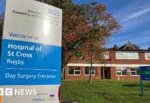 The front of the Hospital of St Cross with a blue and silver NHS sign and brick buildings in the background.