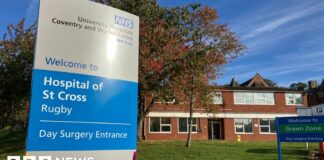 The front of the Hospital of St Cross with a blue and silver NHS sign and brick buildings in the background.