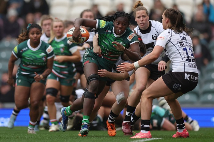 Action-packed rugby scene showing a determined player breaking through tackles, supported by teammates in green jerseys, while the opposing team attempts to stop her