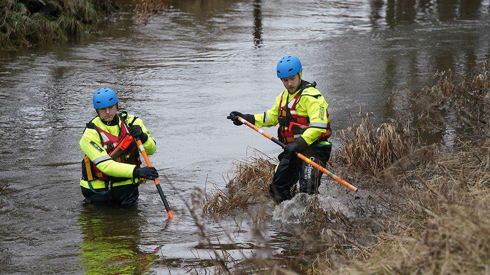 Two men wearing waterproof gear and blue helmets are standing in the river putting poles into it.