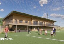 An architect's illustration showing what the new rugby clubhouse would look like. It is a two=-storey building made from wood and has a balcony extending along it. On the pitch are some children being coached, some people standing on the sidelines and a man running.