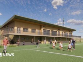 An architect's illustration showing what the new rugby clubhouse would look like. It is a two=-storey building made from wood and has a balcony extending along it. On the pitch are some children being coached, some people standing on the sidelines and a man running.