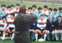 A wax jacket-wearing photographer takes a photo of the South Korean rugby team sat in rows, many wearing white shirts with red and blue hoops on them, and others in blue track suits, while mangers wore blue blazers.