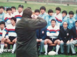 A wax jacket-wearing photographer takes a photo of the South Korean rugby team sat in rows, many wearing white shirts with red and blue hoops on them, and others in blue track suits, while mangers wore blue blazers.