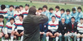 A wax jacket-wearing photographer takes a photo of the South Korean rugby team sat in rows, many wearing white shirts with red and blue hoops on them, and others in blue track suits, while mangers wore blue blazers.