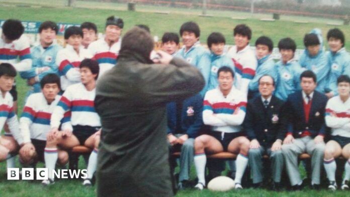 A wax jacket-wearing photographer takes a photo of the South Korean rugby team sat in rows, many wearing white shirts with red and blue hoops on them, and others in blue track suits, while mangers wore blue blazers.