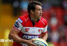 Tom Voyce playing for Gloucester, wearing a red and white shirt and carrying a ball.