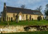 A small church with a white door. It sits in a graveyard, in a raised area with a wall in the foreground of the image.