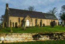 A small church with a white door. It sits in a graveyard, in a raised area with a wall in the foreground of the image.